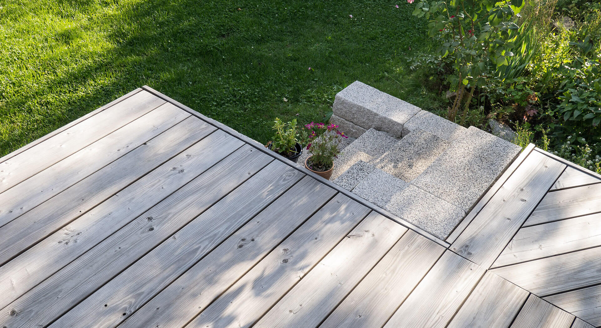 Terrasse im Sommer, gebaut aus Alpin Terrassendielen. Die Terrassendielen sind gleichmäßig vergraut. Im Hintergrund ein Rasen und einige Pflanzen.